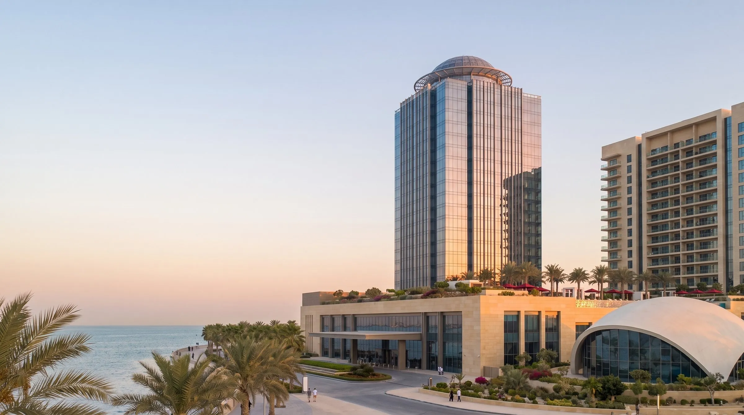 A 36-floor glass hotel tower with a domed crown, a residences block, a pool deck and a shell-roofed ballroom on a seafront promenade at sunset.